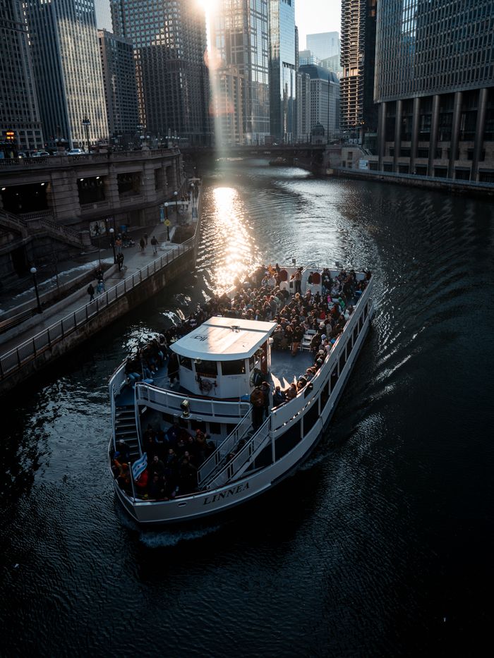 Tour boat filled with passengers travels along the Chicago River with a stunning cityscape view at sunset.